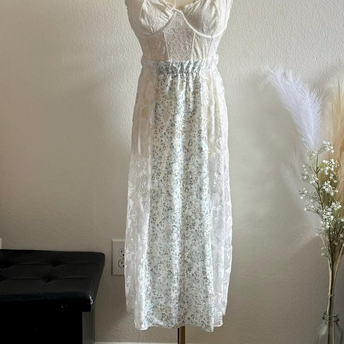 White lace dress on a mannequin against a plain wall with a black bench and decorative plants in the background.